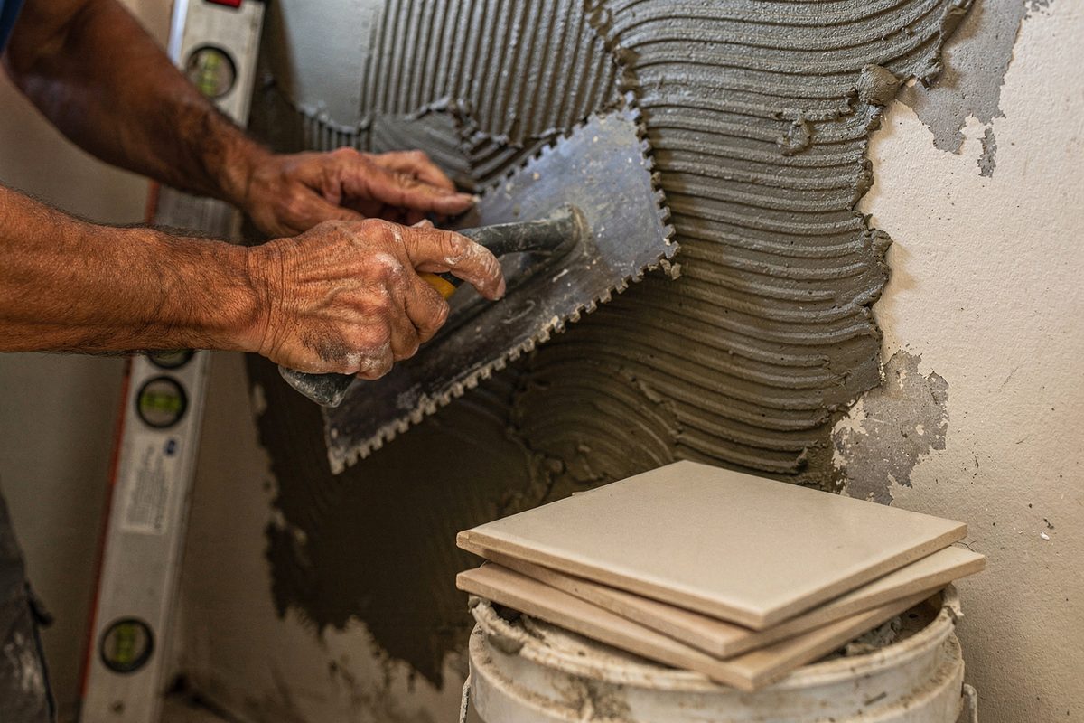 Hands spreading tile adhesive on a bathroom wall with a notched trowel